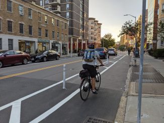 A person riding a bike in a curbside bike lane, separate from the general traffic lane by a painted buffer and plastic flexpost.