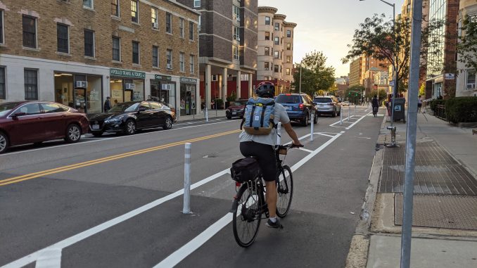 A person riding a bike in a curbside bike lane, separate from the general traffic lane by a painted buffer and plastic flexpost.