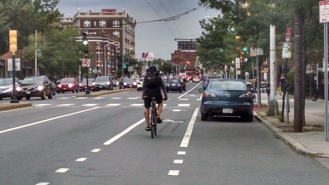 A view of Mass Ave. There are 4 general travel lanes, separated by a median. A cyclist rides in a paint-only bike lane between the general travel lane and a curbside parking lane.