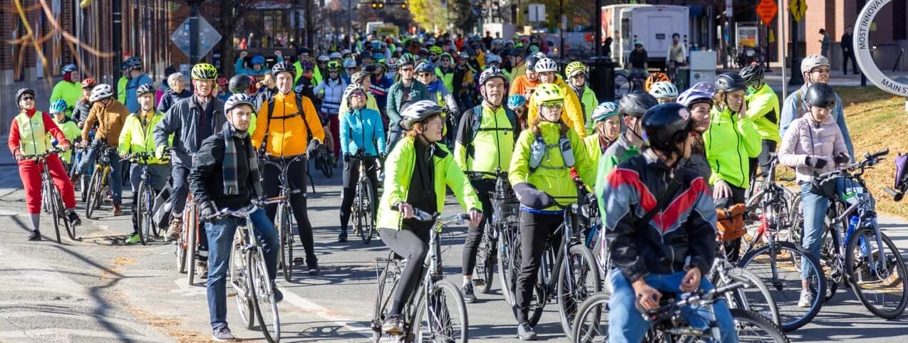 A large group of bicyclists, many wearing reflective gear, waiting on their bikes in Cambridge.